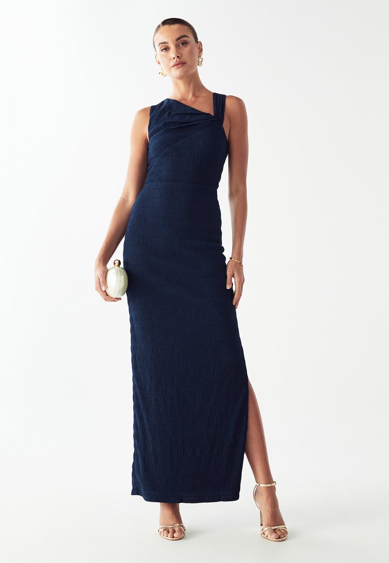 Robe maxi texturée bleu marine avec une bretelle, détails froncés et fente latérale haute. Associée avec des talons dorés et une pochette.