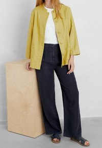 Giacca gialla in lino con scollatura rotonda e bottoni frontali, abbinata a pantaloni larghi scuri. Sandali casual completano l'outfit.