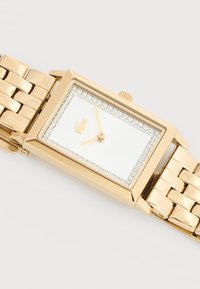 Ορθογώνιο χρυσό ρολόι χειρός με λευκό καντράν, περίγραμμα από κρύσταλλο, χρυστά δείκτες και μικρό χρυσό λογότυπο σκίουρου σε συνδεδεμένο χρυσό βραχιόλι.