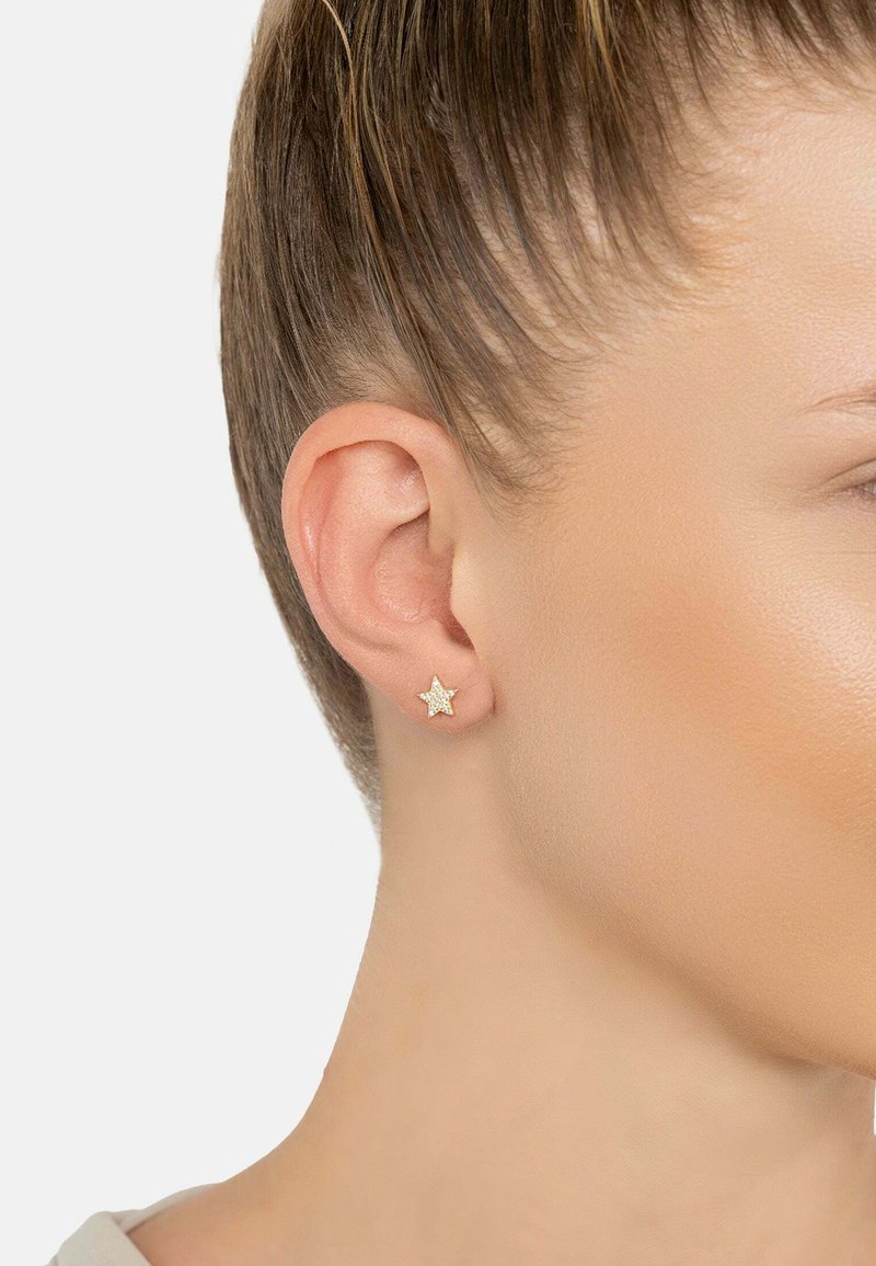 Close-up of a woman’s ear with light brown hair tied back, wearing a small gold star-shaped earring with embedded crystals.