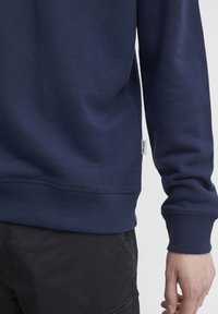 Tamsiai mėlynas džemperis, pagamintas iš medvilnės mišinio, su rūbų apykaklėmis ir apačia, šone yra nedidelis audinio etiketė.