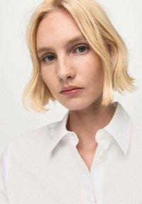 Biela košeľa na gombíky s priliehavým golierom, ktorú nosí osoba s krátkymi blond vlasmi a svetlými pehami, na neutrálnom pozadí.