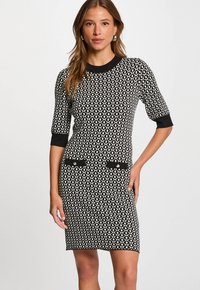 Frau trägt ein knielanges Kleid mit schwarz-weißen geometrischen Mustern, das bis zu den Ellbogen reichende Ärmel hat und schwarze Abschlüsse sowie Taschendetails aufweist.