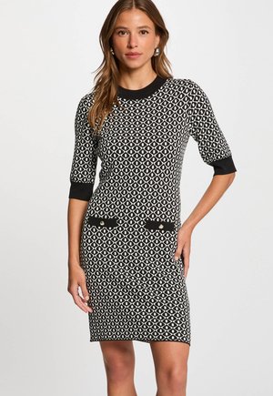 Woman wearing a black and white geometric patterned knee-length dress with elbow-length sleeves and black cuff and pocket details.