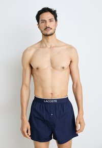 Navy blue cotton boxers with an elastic waistband featuring a white Lacoste logo, front button closure, and smooth texture.