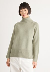 Woman with short black hair wearing a light green turtleneck sweater and white pants against a plain white background.