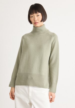 Woman with short black hair wearing a light green turtleneck sweater and white pants against a plain white background.