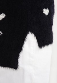 Black fuzzy sweater with a heart pattern in white. Features a cropped design with a scalloped hem. White pants visible underneath.