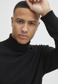 Zwarte coltrui met een geribbelde kraag en mouwen, met een gladde, slim fit ontwerp. Het model poseert met een hand op zijn voorhoofd.