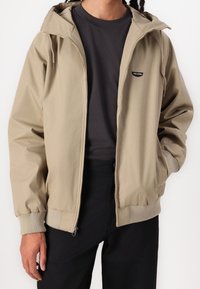 Veste légère beige avec capuche, poignets élastiques, fermeture éclair frontale et écusson logo. Portée par-dessus une chemise foncée, associée à un pantalon noir.