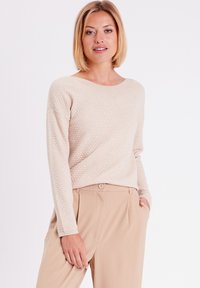 Beige strukturierter Pullover mit einem Bootsausschnitt und langen Ärmeln, kombiniert mit hellbraunen Hosen mit Knopfverschluss und seitlichen Taschen.
