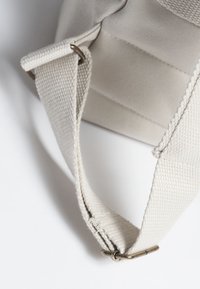 Canvasväska rem i ljusbeige med en texturerad yta, metalldetaljer och en vikt design vid fästpunkten.