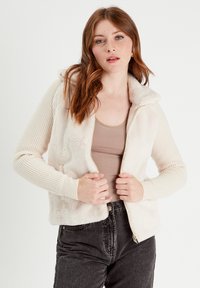 Chaqueta con cremallera de mezcla de pelaje sintético y punto en color crema, con un suave cuello y mangas acanaladas, combinada con una camiseta sin mangas beige y jeans oscuros.