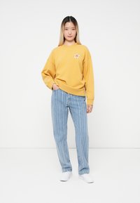 Ungdom som står med en hånd i lommen, iført en sennepsfarget genser med blomstermotiv, stripete blå jeans og hvite joggesko.