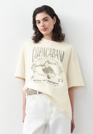 Giovane donna con capelli ricci e scuri che indossa una maglietta oversize color crema con la scritta "Copacabana" e una grafica di tucano, abbinata a pantaloni bianchi.