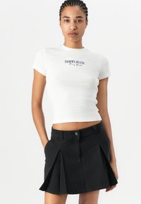 Top court côtelé blanc avec le logo bleu "TOMMY JEANS New York", associé à une jupe mini plissée noire avec poches et fermeture à bouton.