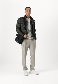 Dark green quilted jacket with four front pockets, gray graphic sweater, cream trousers, and white sneakers, holding a small black shoulder bag.