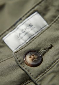 Olivgrön tyg med en stor brun knapp och en vit etikett med texten "JACK'S." Synliga sömnadsdetaljer och en strukturerad yta.