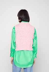 Henkilö, jolla on lyhyet tummat hiukset, pukeutunut vaaleanpunaiseen neulottuun hihattomaan liiviin kirkkaan vihreän pitkähihainen paidan ja vaaleansiniset farkut päällä, kääntyi pois.