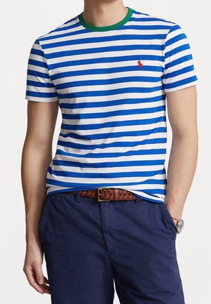 Mężczyzna w niebiesko-białej pasiastym t-shircie z zielonym kołnierzykiem, granatowych spodniach, brązowym plecionym pasku i zegarku na rękę.