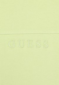 Tejido ajustado de color verde claro con una textura suave. Presenta un logo bordado de "GUESS" en un tono ligeramente más oscuro, centrado en la prenda.