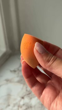 Hand holding a small orange makeup sponge with a teardrop shape and embossed brand logo.