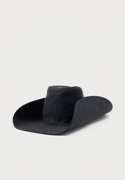 Μαύρο καουμπόικο καπέλο με φαρδιά γείσο και ψηλό, στρογγυλεμένο στέμμα, τοποθετημένο σε απλή λευκή επιφάνεια.