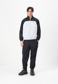 Jachetă de vânt neagră și gri, cu manșete elastice, fermoar frontal și logo. Asortată cu pantaloni negri, lejeri și adidași întunecați, texturați.