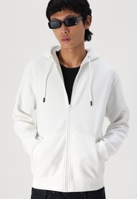 Haut à fermeture éclair blanc en tissu doux, avec une poche frontale, une capuche ajustable à cordon de serrage et des poignets côtelés. Porté par-dessus un t-shirt noir.