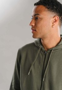 Olivenfarvet zip-up hoodie med snoretræk, der har et tekstureret stof og metal detaljer på lynlåsen. Klar, blød og afslappet design.