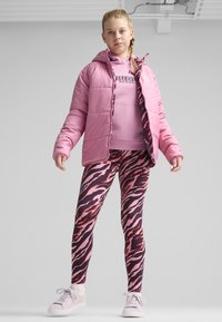 Lyserød polstret jakke med hætte, lagdelt over en lyserød sweatshirt. Sorte og lyserøde zebra-mønstrede leggings. Hvide sneakers fuldender outfittet.