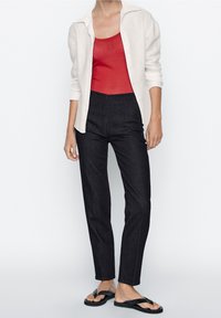 Zwarte denim hoge taille broek, ribbelige rode tanktop en een witte button-up cardigan. Zwarte slippers maken de outfit compleet.