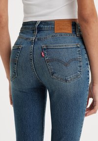 Jeans de mezclilla azul con cintura alta, que cuentan con bolsillos traseros y una etiqueta de cuero marrón con el logo de la marca. Textura suave, ligero desgaste.