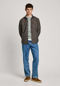 Bruin geruit overhemd over een lichtgroene sweater, gecombineerd met blauwe jeans en bruine schoenen, tegen een effen achtergrond.