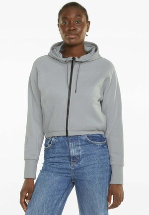 Kobieta w jasnoszarej bluzie na suwak z kapturem i niebieskich jeansach z wysokim stanem, stojąca na białym tle.