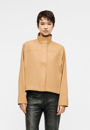 Žena s krátkymi tmavými vlasmi, oblečená v béžovej bunde s vysokým golierom a čiernych štruktúrovaných nohaviciach, stojaca proti jednoliatemu svetlému pozadiu.