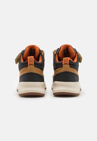 Des sneakers noir et beige présentent un dessus texturé, un intérieur rembourré orange et une semelle en caoutchouc avec un motif de bande de roulement distinct. Fermeture à strap auto-agrippant.