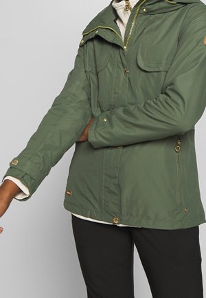 Persoon die een groene jas met gouden knopen draagt en de mouw aanpast, gekleed in een zwarte broek en een witte binnenlaag.