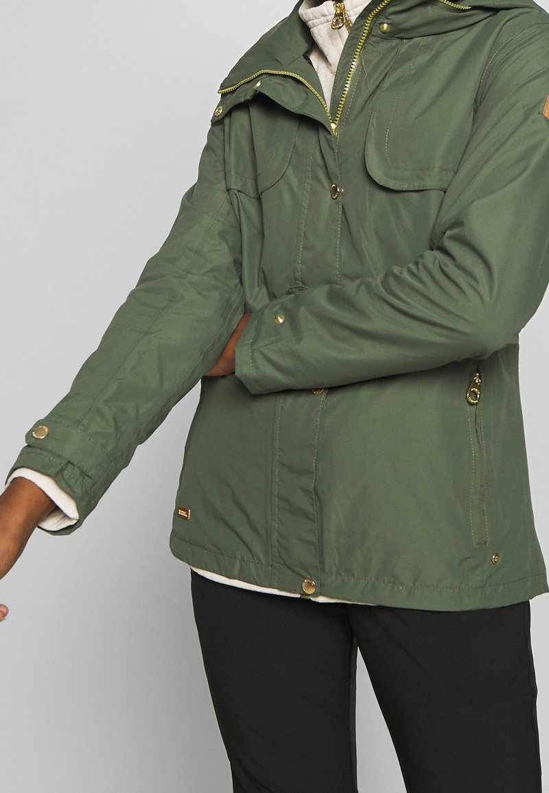 Personne portant une veste verte avec des boutons dorés ajustant la manche, vêtue d’un pantalon noir et d’une couche intérieure blanche.