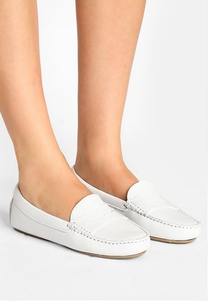 Fødder iført hvide læder penny loafers med syninger og flade tan såler på en hvid baggrund.