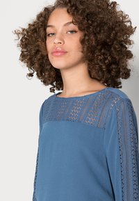 Blusa azul com uma parte superior de renda texturizada e tecido liso. Decote redondo com mangas longas, design drapeado e ligeiro brilho.