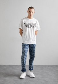 T-shirt de algodão branco com estampagem gráfica preta, combinada com jeans de ganga azul com um padrão de graffiti. Ténis brancos completam o visual.