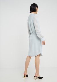 Himmelblaues, gestreiftes, knielanges Kleid mit langen Ärmeln und verstellbarem Taillenbund. Kombiniert mit schwarzen Slippers mit runden goldenen Akzenten.