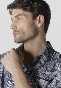 Homme aux cheveux courts et foncés avec une barbe légère, portant une chemise bleue et blanche à motif de feuilles, regardant vers la gauche avec la main près du col.