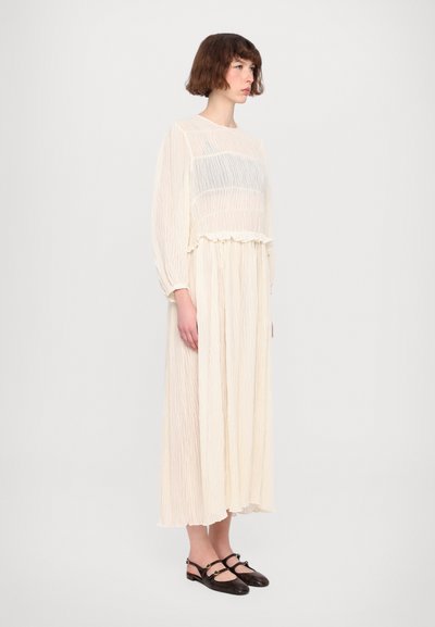 Crinkled cream dress with long sleeves, ruffled waist, and a sheer bodice layer. Paired with dark, textured Mary Jane shoes.