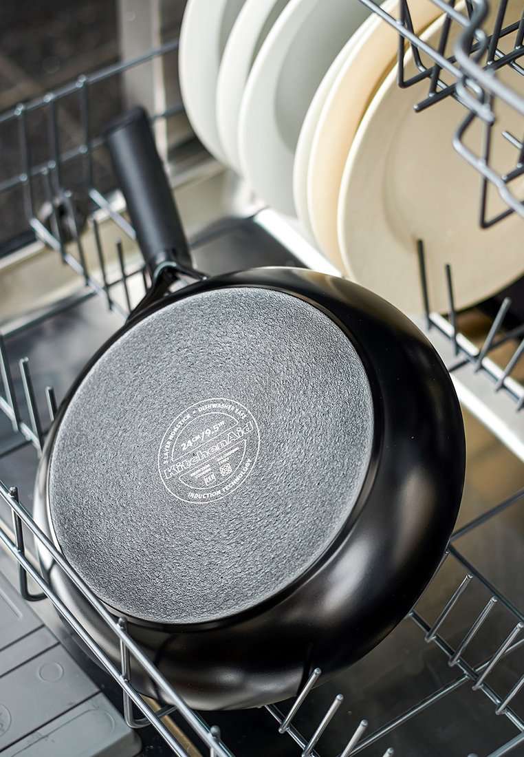 Schwarze Bratpfanne mit strukturierter Oberfläche, mit einem Logo auf der Unterseite, die in einer Spülmaschine neben hellen Tellern liegt.