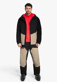 Schwarze und beige Skijacke mit Kapuze, Reißverschluss vorne und Reißverschlusstaschen, kombiniert mit passenden beige und schwarzen Skihosen und dunklen Skistiefeln.