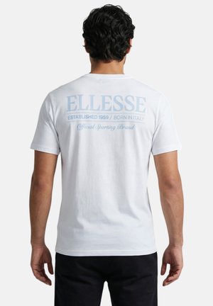 Homme aux cheveux bouclés foncés portant un t-shirt blanc avec un texte bleu clair « ELLESSE Established 1959 / Born in Italy » dans le dos.