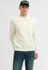 Suéter de punto acanalado color crema con cuello en barco y mangas largas, combinado con jeans de color azul oscuro. Diseño sencillo, textura suave, ideal para el uso diario.