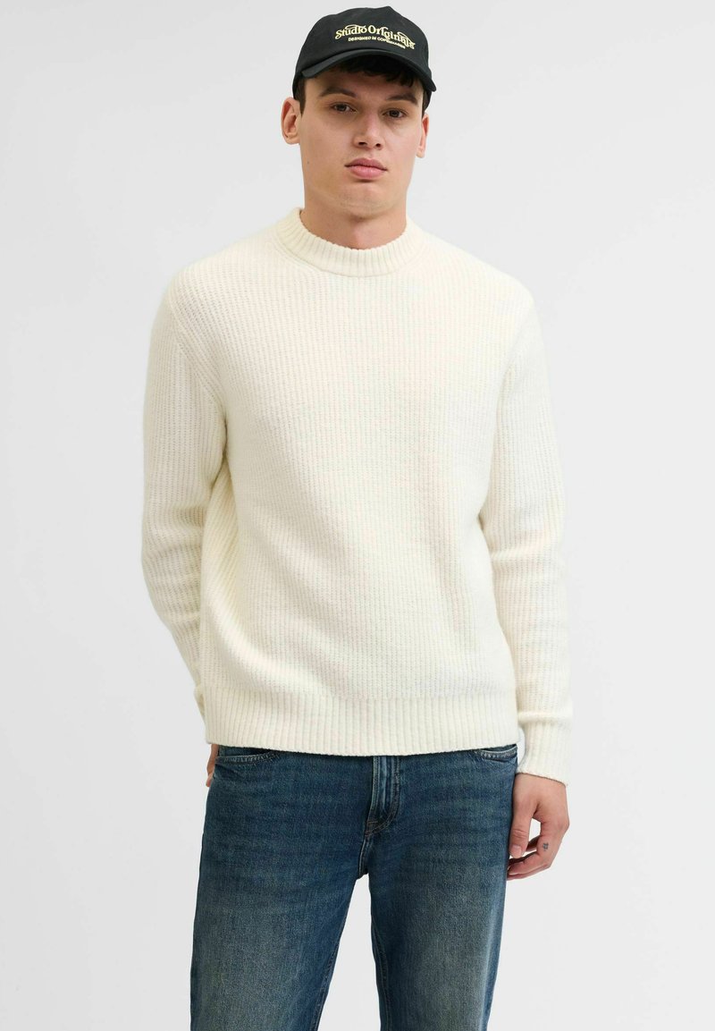 Suéter de punto acanalado color crema con cuello en barco y mangas largas, combinado con jeans de color azul oscuro. Diseño sencillo, textura suave, ideal para el uso diario.